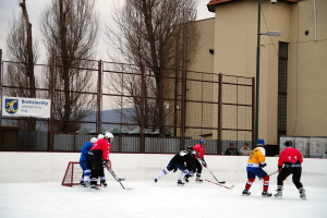 Personen beim Eishockeyspielen auf einem Eisstadion mit Gebäuden, Bäumen, einer Straßenlaterne, einem Namensschild und Zäunen im Hintergrund unter einem Himmel.