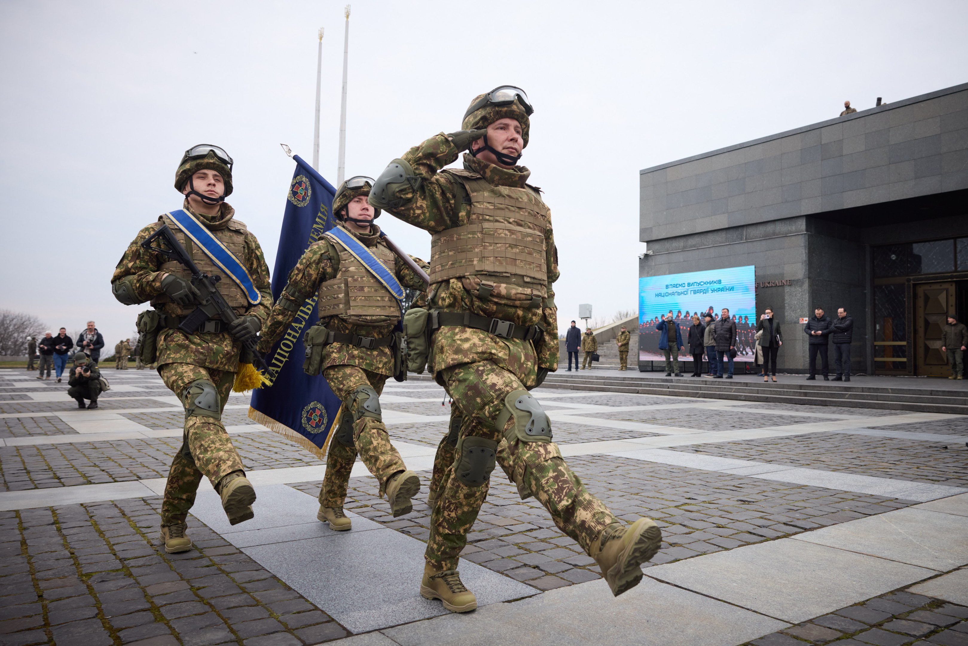 Eine Gruppe ukrainischer Soldaten in Uniform marschiert mit Gewehren und Fahnen die Straße entlang, mit Zivilisten, Bäumen und einem klaren Himmel im Hintergrund.