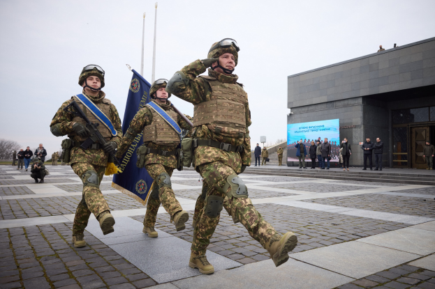 Eine Gruppe ukrainischer Soldaten in Uniform marschiert mit Gewehren und Fahnen die Straße entlang, mit Zivilisten, Bäumen und einem klaren Himmel im Hintergrund.