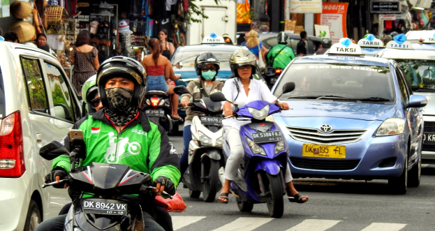 Eine Gruppe von Menschen auf Motorrädern auf einer belebten Straße mit Autos und anderen Fahrzeugen, einige Fahrer tragen Helme, Gebäude, Bäume, Schilder und Geschäfte im Hintergrund.