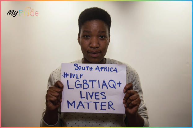 Eine Frau in einem weißen Hemd steht entschlossen vor einer Wand und hält ein Schild hoch, auf dem in fetten schwarzen Buchstaben "LGBTQ+ Lives Matter" steht.