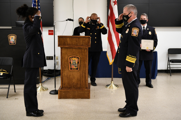 Gruppe uniformierter Personen mit Masken auf einem Podium während einer Vereidigung eines neuen Feuerwehrchefs, der eine Urkunde hält, mit Flaggen, Stühlen, Tischen und einer Wand im Hintergrund.
