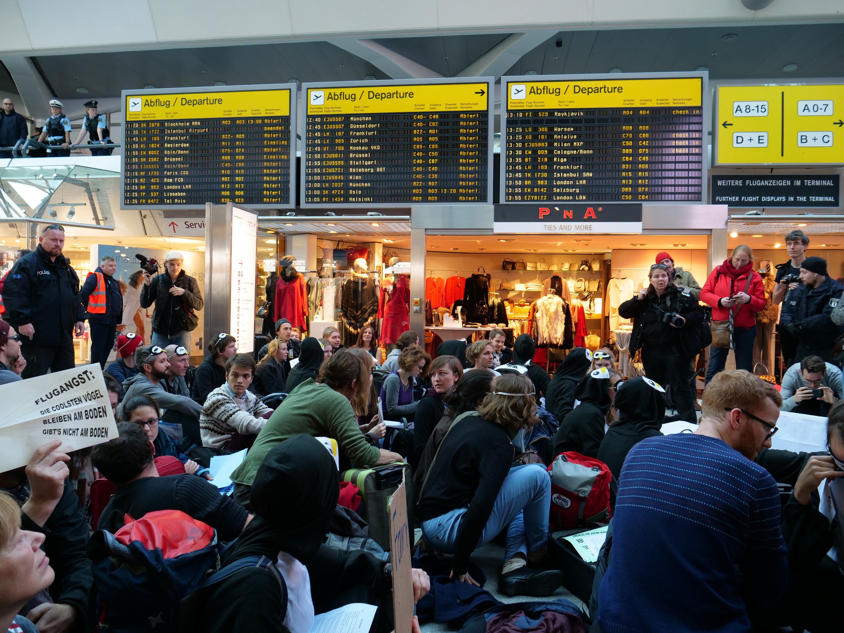 Eine große Gruppe von Menschen in einem Flughafen, einige sitzen mit Taschen und Papieren, andere stehen, mit Texttafeln, Schaufensterpuppen in Kleidern und Deckenbeleuchtung im Hintergrund während einer Demonstration.