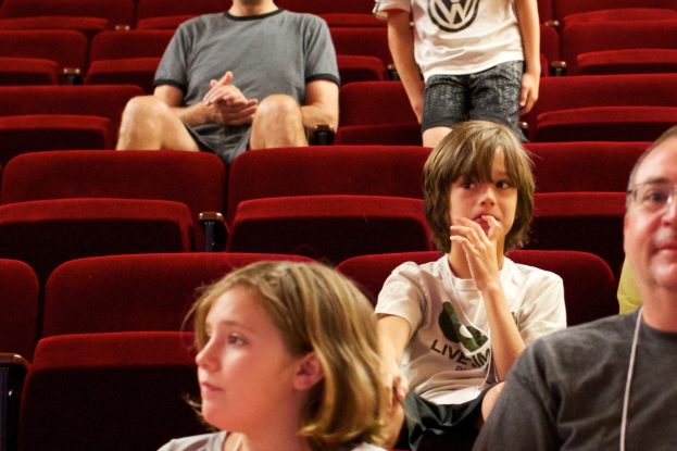Eine Gruppe von Menschen in einem Kino mit roten Stühlen, ein Junge in einem weißen T-Shirt und schwarzen Hosen in der Mitte.