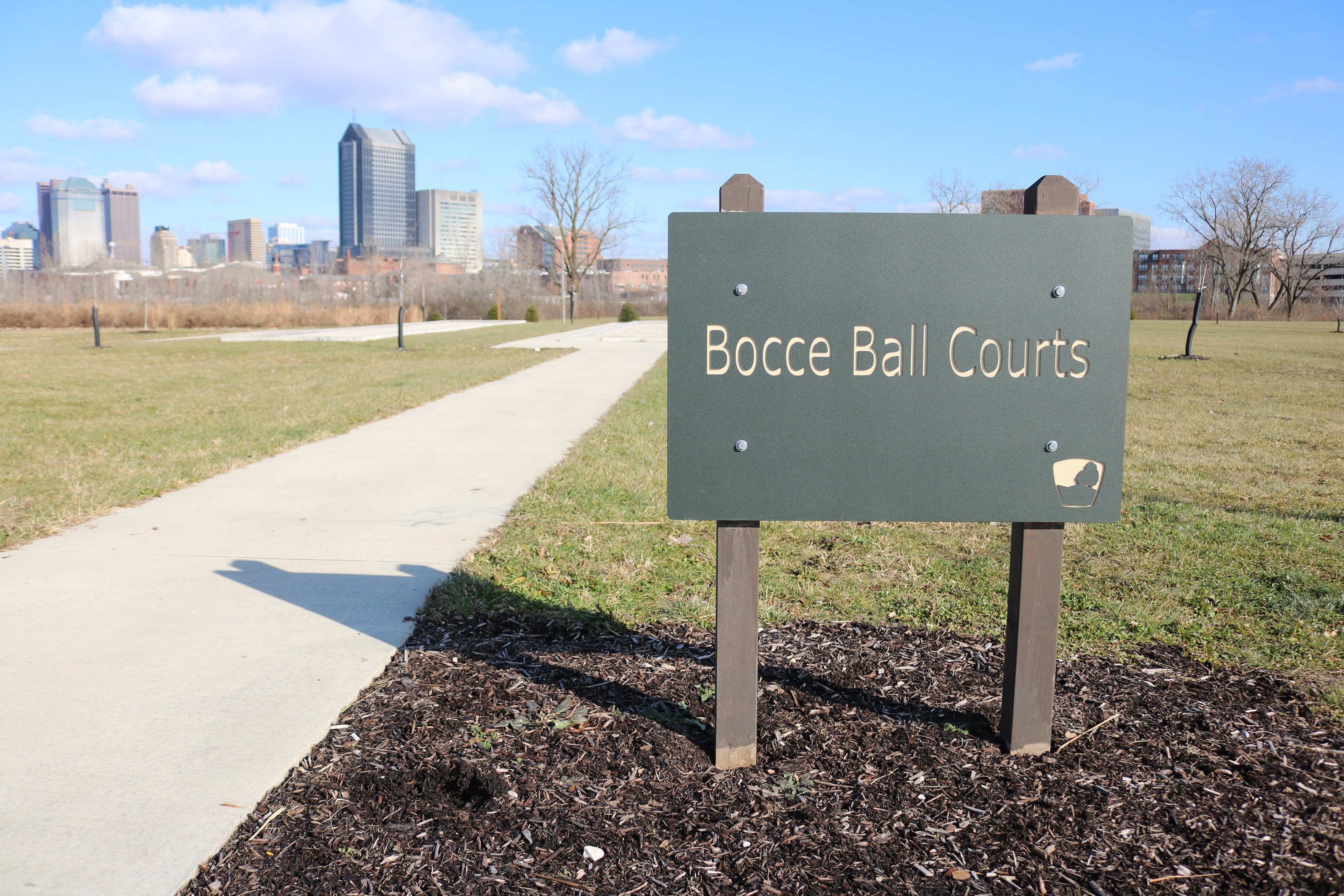 Ein Schild am Boden in der Nähe eines Weges und Bäume trägt die Aufschrift "Bocce Ball Courts in Indianapolis, Indiana" mit einem bewölkten Himmel im Hintergrund.