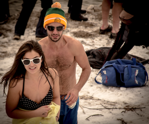 Ein Mädchen steht am Strand und hält ein Handtuch, neben einer Person ohne Hemd, mit mehreren Personen im Hintergrund und einer Tasche unten. Beide, das Mädchen und die Person ohne Hemd, tragen eine Brille.