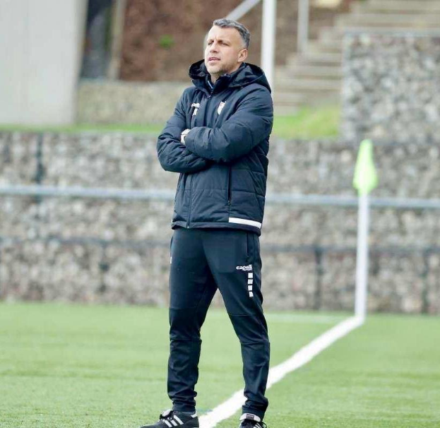 Ein Fussballtrainer, der auf einem Feld steht und die Ärmel überkreuzt, trägt eine schwarze Jacke und Hose, vor einem Zaun, Treppe und Wand während eines Trainings.
