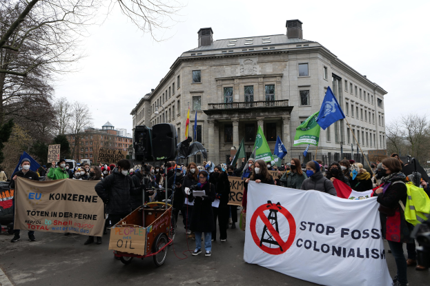 Große Gruppe von Menschen bei einer Protestaktion gegen fossile Brennstoffe, die Schilder und Fahnen tragen, mit einem Fahrzeug im Vordergrund und Gebäuden, Bäumen und einem klaren blauen Himmel im Hintergrund.