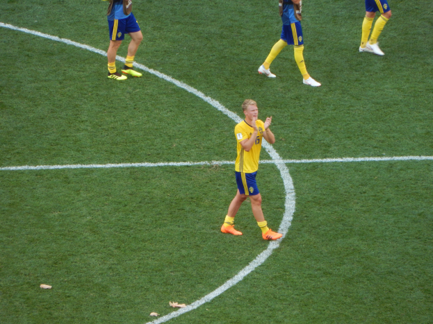 Eine Gruppe von Männern in Fußballtrikots und -schuhen steht auf einem Fußballfeld, wobei eine Person in der Mitte klatscht.