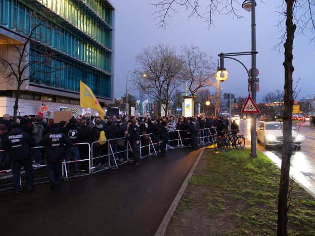 Eine große Gruppe von Menschen steht vor einem Gebäude in Berlin und hält Schilder, mit Barrikaden, Fahrrädern, Laternenmasten, Schildern, Bäumen und Gras auf dem Boden, unter einem sichtbaren Himmel.