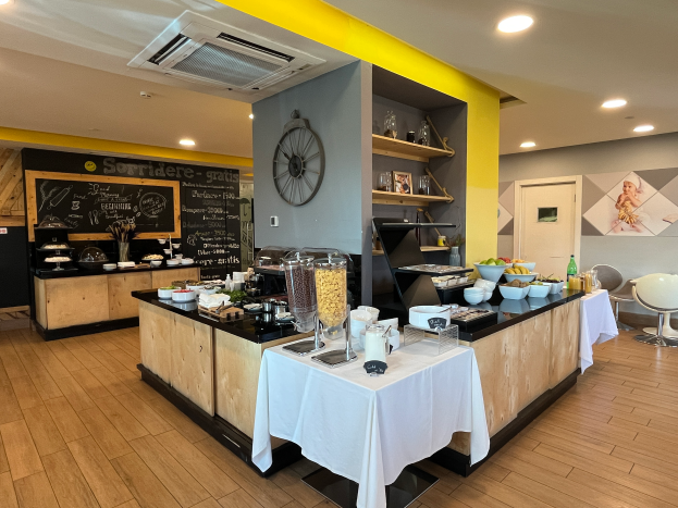 Ein Restaurantinnenraum mit Tischen und Stühlen, Speisen, einer Tafel mit Text, einer Wanduhr, Deckenleuchten, einer Klimaanlage und einer Tür im Hintergrund, Located in the Hotel de Ville in Paris.