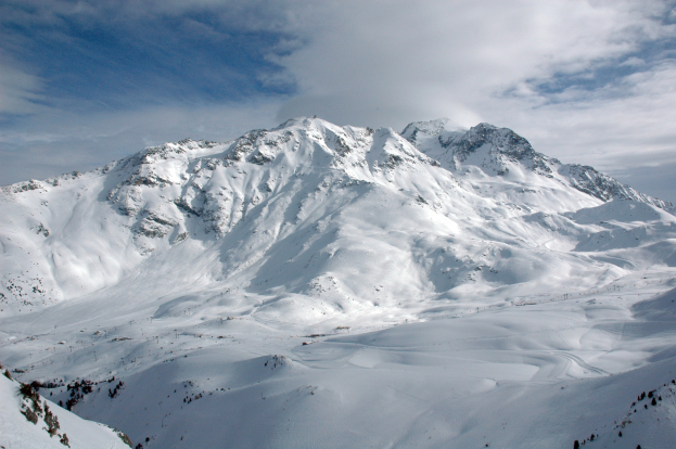 Ein schneebedeckter Berg mit einigen Skifahrern, die darauf hinabfahren, umgeben von einer bewölkten Himmel und unberührtem Schnee.