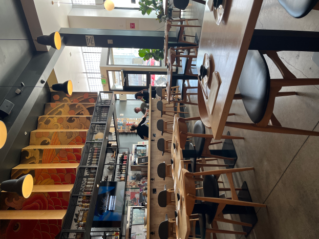 Ein Restaurantinnenraum mit Tischen und Stühlen in Reihen, einige mit Tellern und Gläsern, Menschen stehen herum, Treppe auf der linken Seite, hängende Lichter und eine Pflanze im Hintergrund.