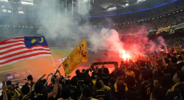 Eine große Menschenmenge in einem Stadion mit brennenden Fackeln in der Luft, einige halten Fahnen und eine Person hält eine Kamera, mit Rauch im Hintergrund und Schildern, die "Malaysia vs Malaysia Live Streaming" anzeigen.