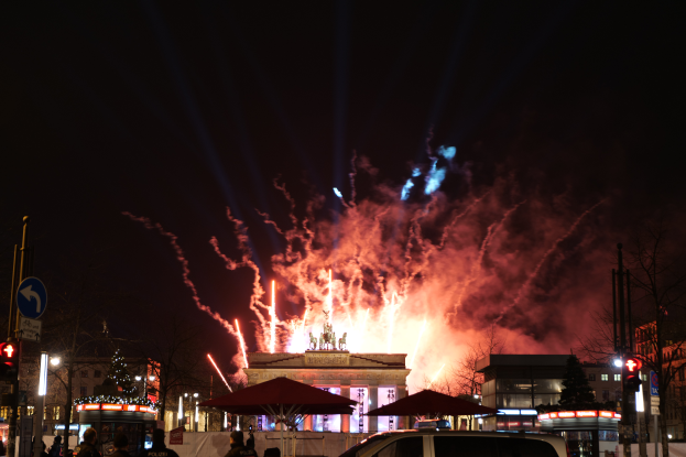 Ein Auto steht vor dem Brandenburger Tor in Berlin, Deutschland, mit Feuerwerk, das den Nachthimmel erhellt, Menschen mit Schirmen drumherum und Gebäude und Bäume im Hintergrund.