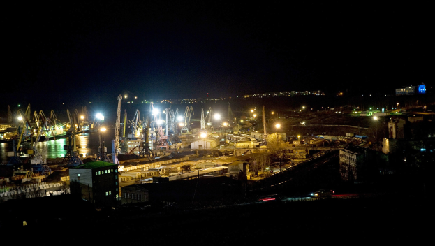 Eine nächtliche Stadtansicht mit beleuchteten Gebäuden, fahrenden Fahrzeugen auf Straßen und Wasser im Vordergrund vor einem dunklen Himmel.