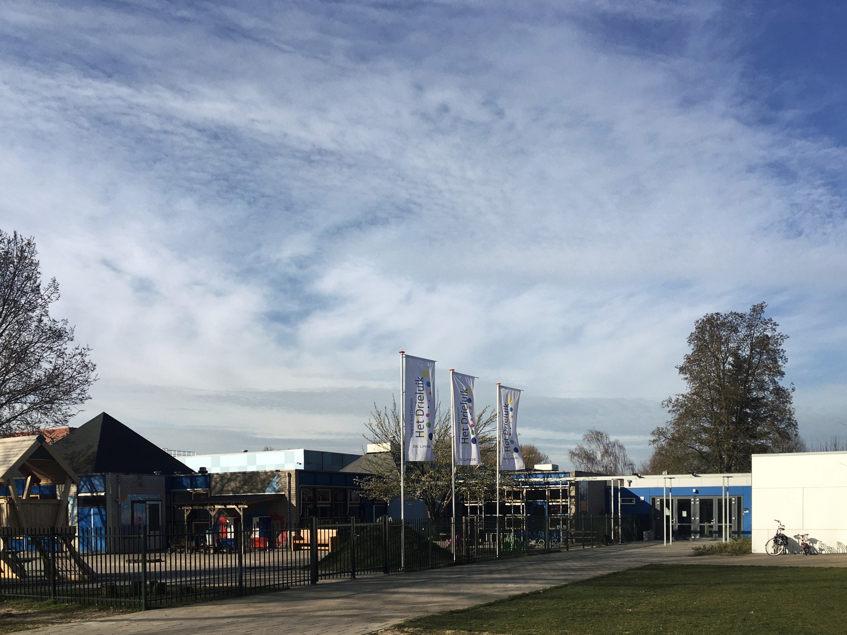 Großes Gebäude mit einem grasigen Bereich davor, umgeben von einem Metallzaun, mit einem Weg, Bäumen, Pfosten, Bannern, einem geparkten Fahrrad, nahen Häusern und einem bewölkten Himmel, als neuer Spielplatz der Schule identifiziert.
