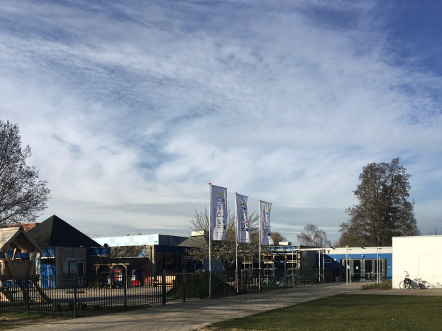 Großes Gebäude mit einem grasigen Bereich davor, umgeben von einem Metallzaun, mit einem Weg, Bäumen, Pfosten, Bannern, einem geparkten Fahrrad, nahen Häusern und einem bewölkten Himmel, als neuer Spielplatz der Schule identifiziert.