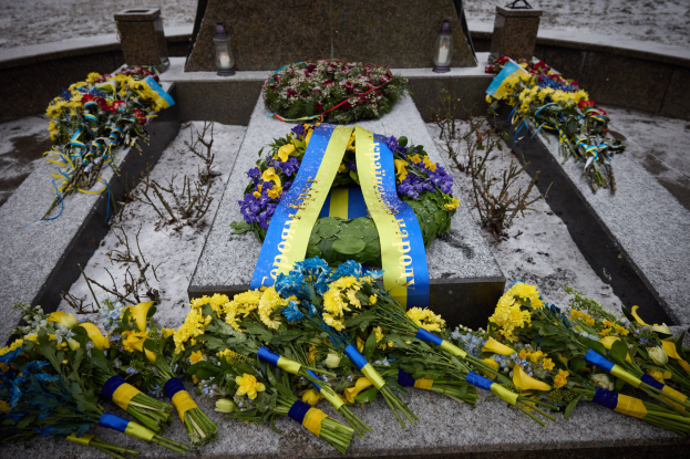 Gedenkstätte in Kiew, Ukraine, mit einem Blumenkranz auf einem Grab, das mit einer blauen Schleife markiert ist und von zusätzlichen Blumensträußen umgeben ist, vor einem verschneiten Hintergrund.