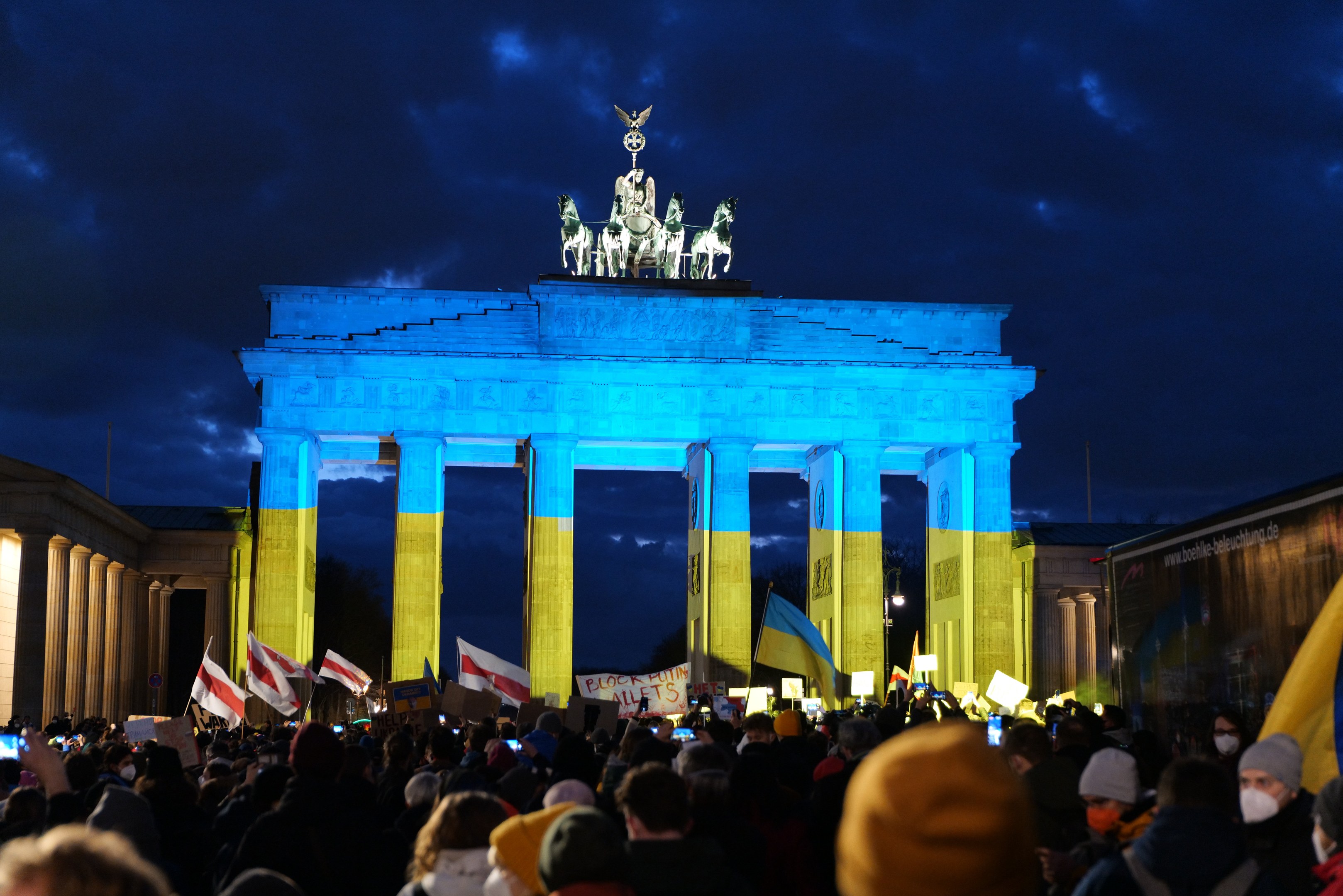 Menschenmenge mit Fahnen und Schildern vor dem Brandenburger Tor in Berlin, mit einer Fahne auf der rechten Seite des Bildes.