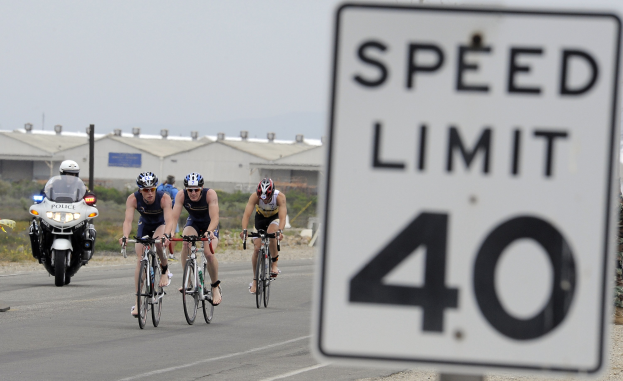 Gruppe von Menschen mit Helmen auf Fahrrädern die Straße entlang fahren neben einem Geschwindigkeitsbegrenzungsschild, mit Gebäuden, Masten, Pflanzen, Bäumen und einem klaren blauen Himmel im Hintergrund.