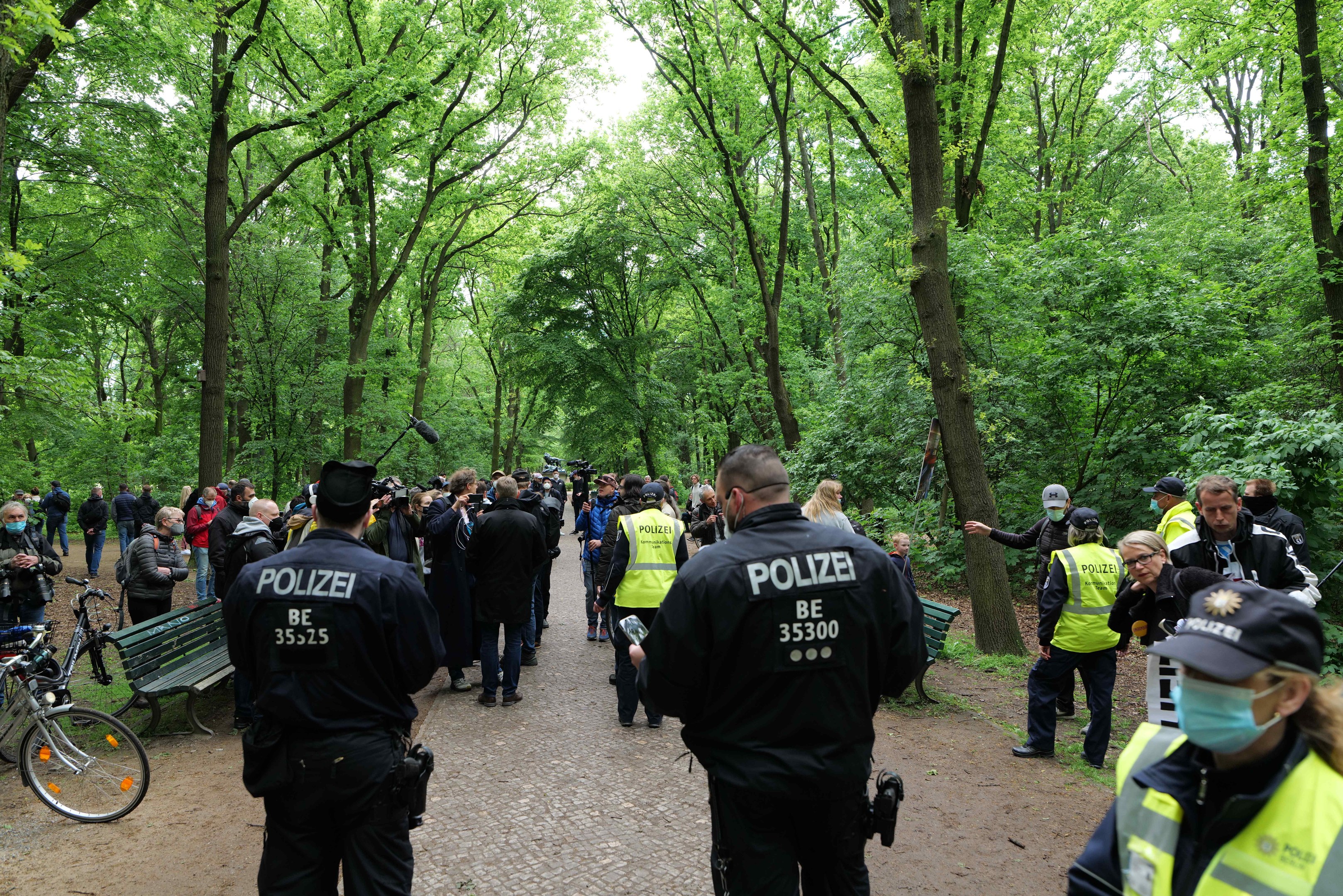 Polizisten vor einer Menge bei einer Demonstration im Freien, mit Fahrrädern und einer Bank im Vordergrund und Bäumen im Hintergrund.