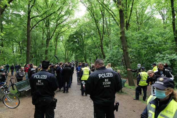 Polizisten vor einer Menge bei einer Demonstration im Freien, mit Fahrrädern und einer Bank im Vordergrund und Bäumen im Hintergrund.