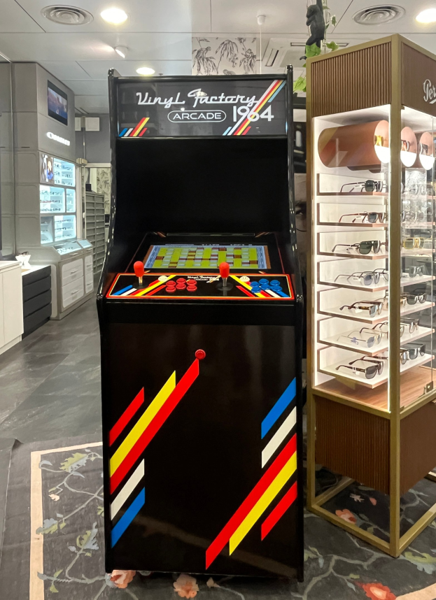 Ein Geschäft mit einem zentralen Retro-Arcade-Automaten, einer rechten Stange mit Gläsern und Hintergrund-Elementen wie Schränken, einem Fernseher und anderen Gegenständen, mit Deckenlampen, die eine Retro-Atmosphäre schaffen.