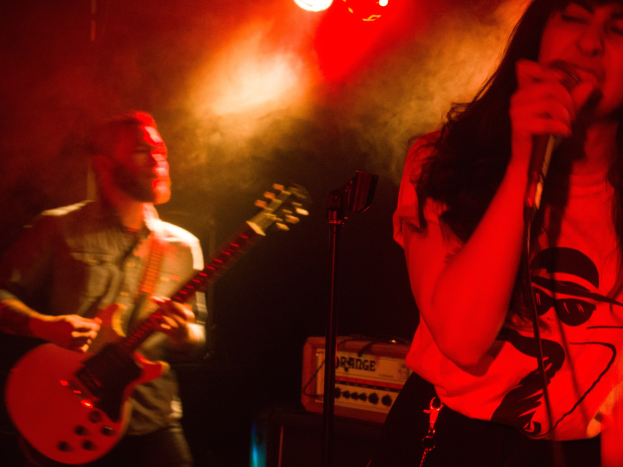 Eine Frau singt in ein Mikrofon auf der Bühne, während ein Mann im Hintergrund eine akustische Gitarre spielt, mit Deckenleuchten, die die Szenerie erhellen.