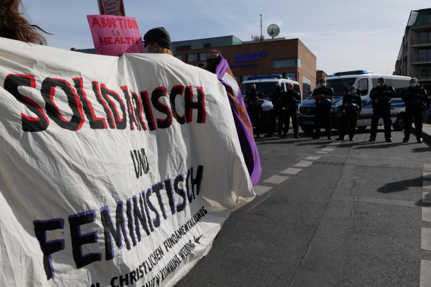 Eine Gruppe von Menschen, die eine Fahne mit der Aufschrift "Solidarität und Feminismus" schwenken, geht eine Straße entlang mit parkenden Fahrzeugen und Gebäuden im Hintergrund unter einem klaren blauen Himmel.