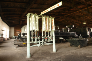Ein Bahnhof mit einem Zug auf den Gleisen, einem Rollstuhl auf dem Boden, einer Signalbox in der Mitte, einem Pfahl mit einer Tafel, einer Wand auf der linken Seite und einer Decke oben.