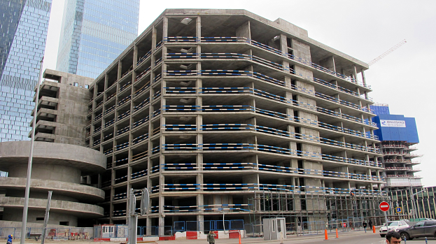 Großer Bau in einer städtischen Umgebung mit Wolkenkratzern, Fahrzeugen, Fußgängern, Verkehrsinfrastruktur und Kränen vor einem klaren Himmel.