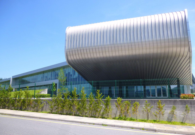 Das neue Hauptquartier der University of Toronto, ein großes Gebäude mit gewölbtem Dach, umgeben von Pflanzen, Gras, einem Weg, einem Zaun, einer Baumgruppe und unter einem bewölkten Himmel.