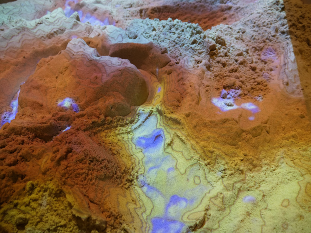 Nahaufnahme einer detaillierten 3D-Karte einer Wüstenlandschaft, die sandfarbene Berge, Täler und Grate zeigt, die von hellem Licht beleuchtet werden.