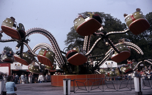 Vergnügungspark-Achterbahn mit sitzenden Menschen, umgeben von einem Zaun und einem Mülleimer, vor Bäumen und einem klaren blauen Himmel.
