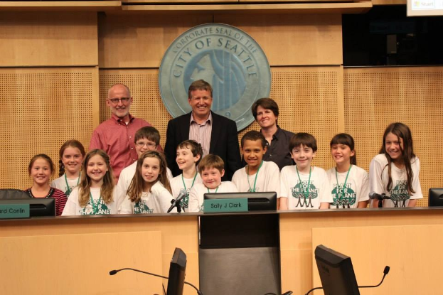 Gruppe von Kindern und Erwachsenen, die vor einem Rednerpult auf einer Stadtratssitzung lächeln, mit Monitoren und Mikrofonen auf einem Tisch im Vordergrund und einem Logo und einem Bildschirm im Hintergrund.