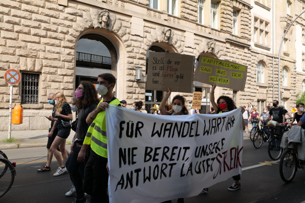 Eine Gruppe von Menschen nimmt an einer Protestaktion gegen die Anti-Kriegsbewegung in Berlin teil, geht eine Straße entlang und hält Schilder und Plakate hoch, während einige Fahrräder fahren, vor einem Gebäude mit Fenstern, Bögen, Säulen und Skulpturen und umgeben von Bäumen und einem Laternenmast.