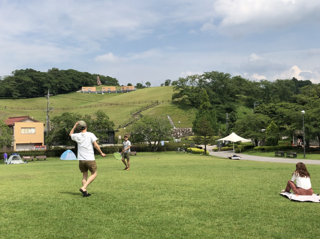 Gruppe von Menschen, die Badminton in einem Park spielt, mit einem Mann, der einen Schläger hält und auf einer Decke auf dem Gras sitzt, in der Nähe von Zelten und Gebäuden, mit Hügeln und bewölktem Himmel im Hintergrund.
