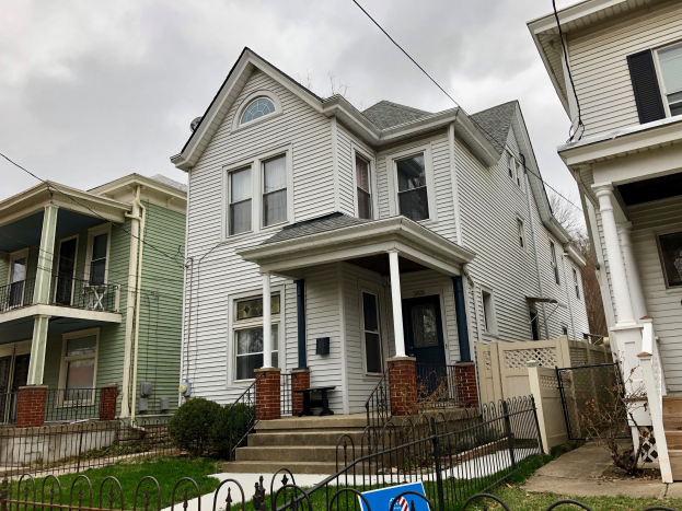 Eine Reihe von Häusern mit Fenstern, Türen, Säulen, Geländern, Stufen und Dächern, umgeben von einem Metallzaun, Gras, Pflanzen, Bäumen und einem bewölkten Himmel, mit einem Mietschild unten, das die Adresse als 5901 S Elmwood Ave, Chicago 60618 angibt.