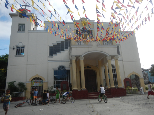 Eine Gruppe von Menschen, die Fahrräder vor einer weißen Kirche mit architektonischen Details, Pflanzen, einem Zaun, einer Fahne, Bäumen und einem bewölkten Himmel fahren, mit bunten Fahnen, die an einem Seil befestigt sind.