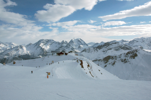Eine Gruppe von Menschen, die eine schneebedeckte Piste mit Bergen im Hintergrund hinabskifahren, unter einem bewölkten Himmel mit glitzerndem Schnee.