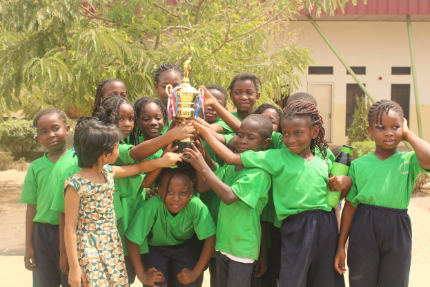 Eine Gruppe von Kindern, die zusammenstehen und eine Trophäe in einer Schulumgebung mit Bäumen und einem Gebäude im Hintergrund halten.
