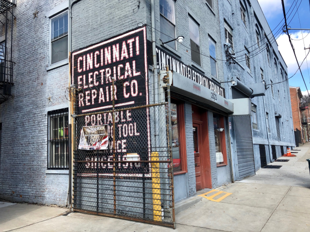 Cincinnati Electrical Repair Co. Gebäude mit einer Tafel, Metallzaun, Mülltonne, Versorgungsmasten, Drähte, Bäume und einem bewölkten Himmel.