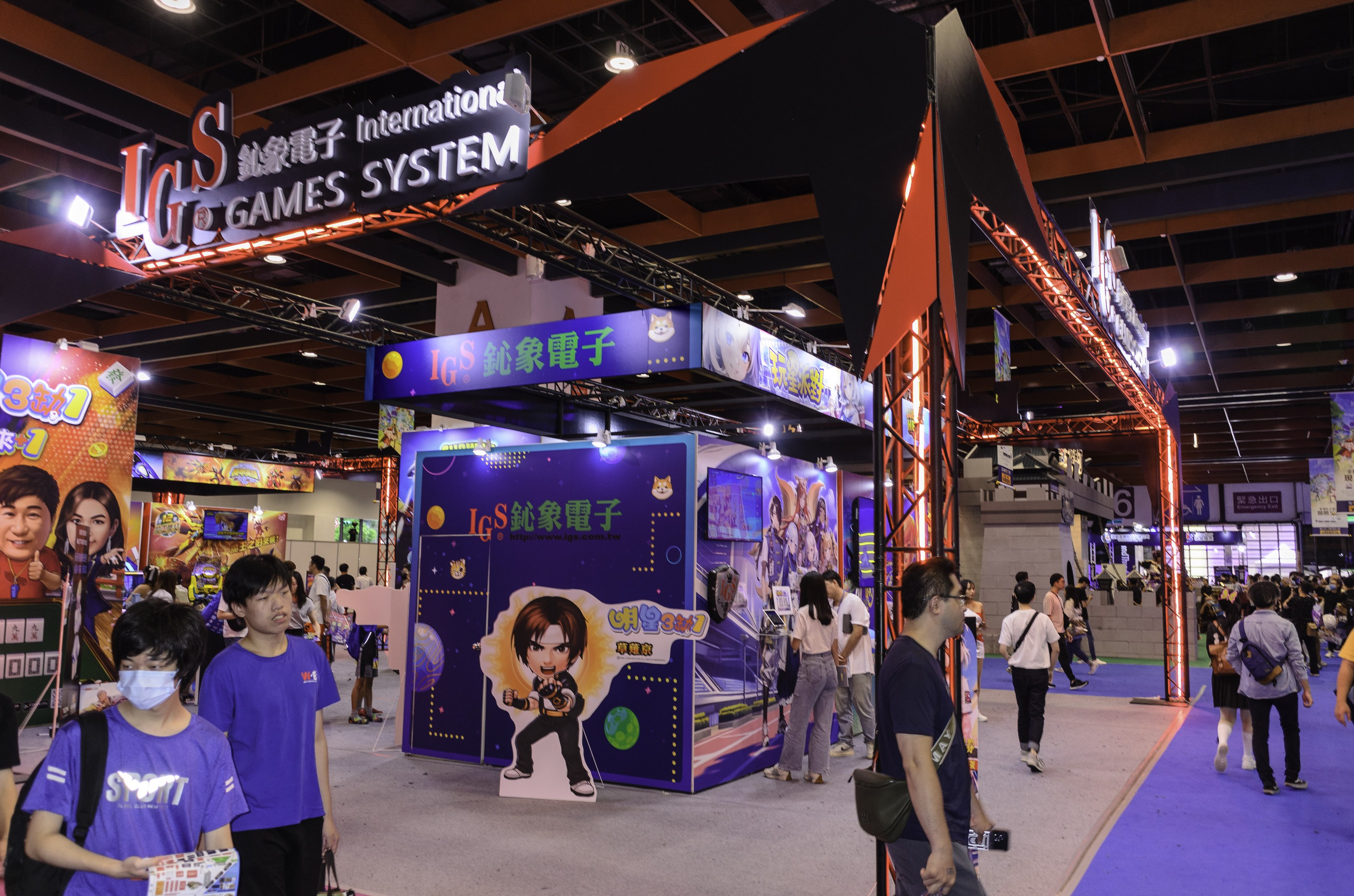 Eine Gruppe von Teilnehmern geht durch einen Konferenzsaal auf einer Spielemesse in China, einige tragen Taschen und halten Gegenstände, mit Displaytafeln, Deckenbeleuchtung und einem Teppichboden.