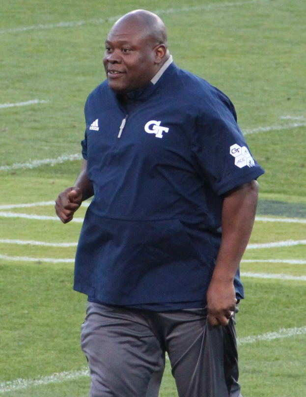 Ein Mann in einem blauen Hemd und grauen Hosen steht auf einem grünen Feld und lächelt mit einem selbstbewussten und entschlossenen Gesichtsausdruck, wahrscheinlich ein Fußballtrainer.