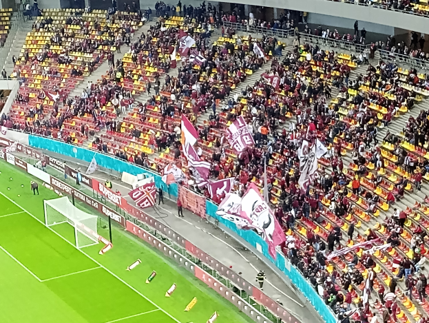 Ein großes Stadion voller Zuschauer, die ein Fußballspiel beobachten, mit einem Tor und Netz auf der linken Seite, einigen Menschen, die stehen und sitzen, sowie Tafeln mit Text, Treppen, Geländern und einer Wand im Hintergrund.