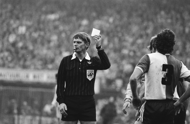 Schwarzes und weißes Bild eines Schiedsrichters, der eine rote Karte vor einer Menge von Menschen in Fußballuniformen hält, mit einem Zaun im Hintergrund.