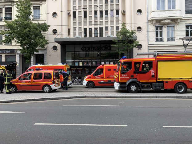 Eine Gruppe von Feuerwehrautos steht auf einer Straße in Paris, mit Menschen auf dem nahen Bürgersteig, Gebäuden, Bäumen und einem Fahrrad im Hintergrund.