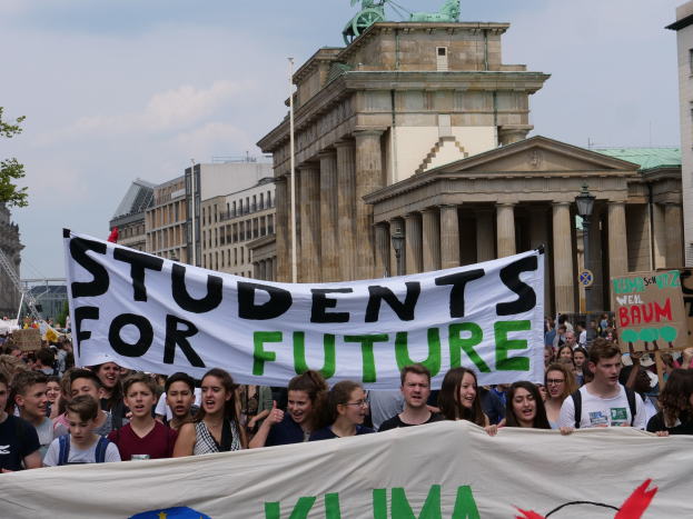 Gruppe von Schülern marschiert in Berlin mit einem leuchtend bunten 'Students for Future'-Schild vor einer Kulisse aus Gebäuden, Bäumen und Himmel.