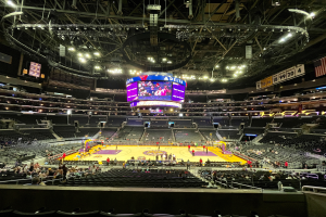 Eine große Arena, das Staples Center, voller Zuschauer bei einem Basketballspiel mit einem großen Bildschirm oben, einer Flagge und Anzeigetafeln rechts und einer Decke mit Stangen und Lichtern.